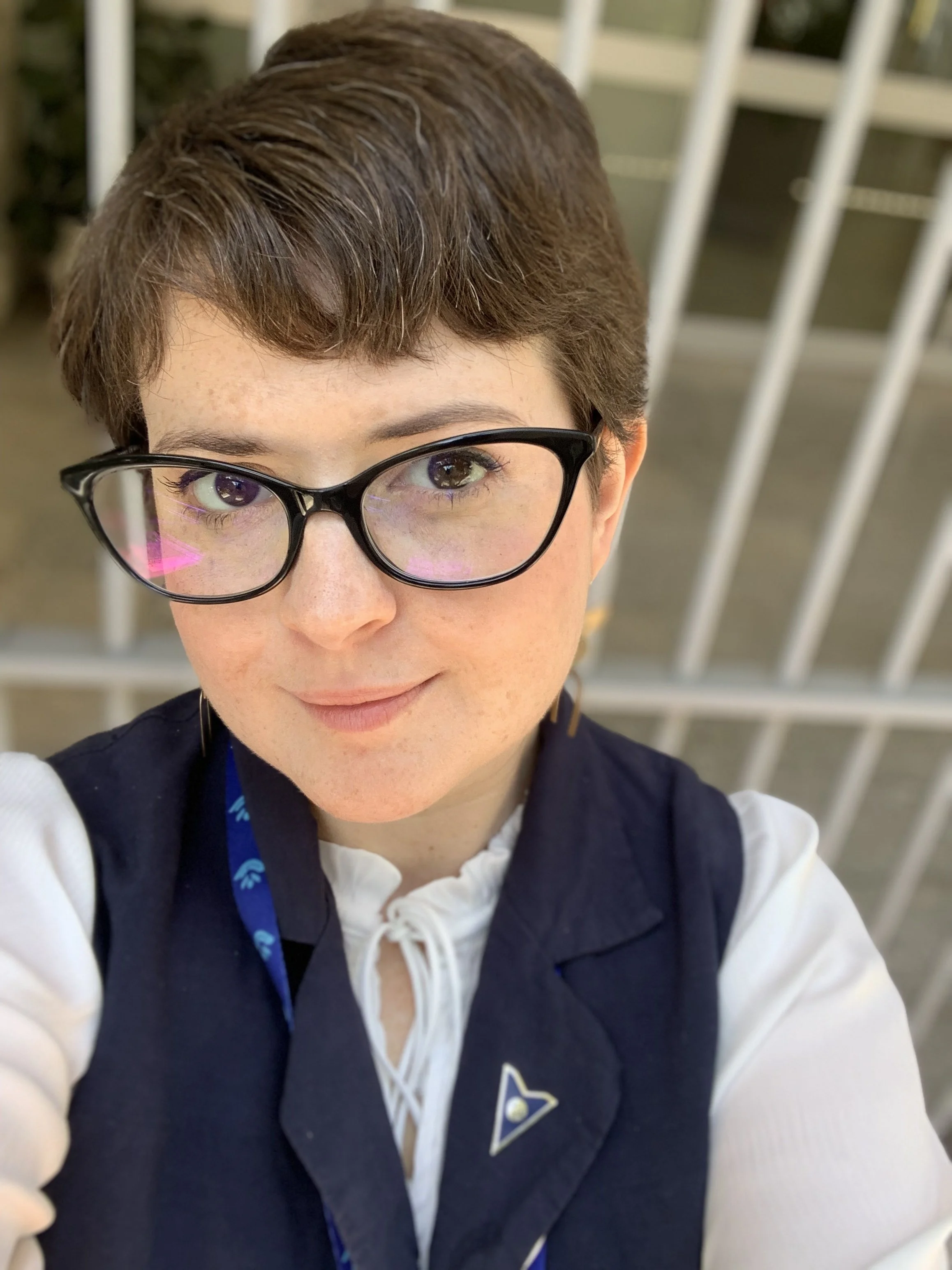 Close-up selfie of a person with short brown hair, wearing black glasses, a white blouse with a tie, and a navy vest, standing outdoors.
