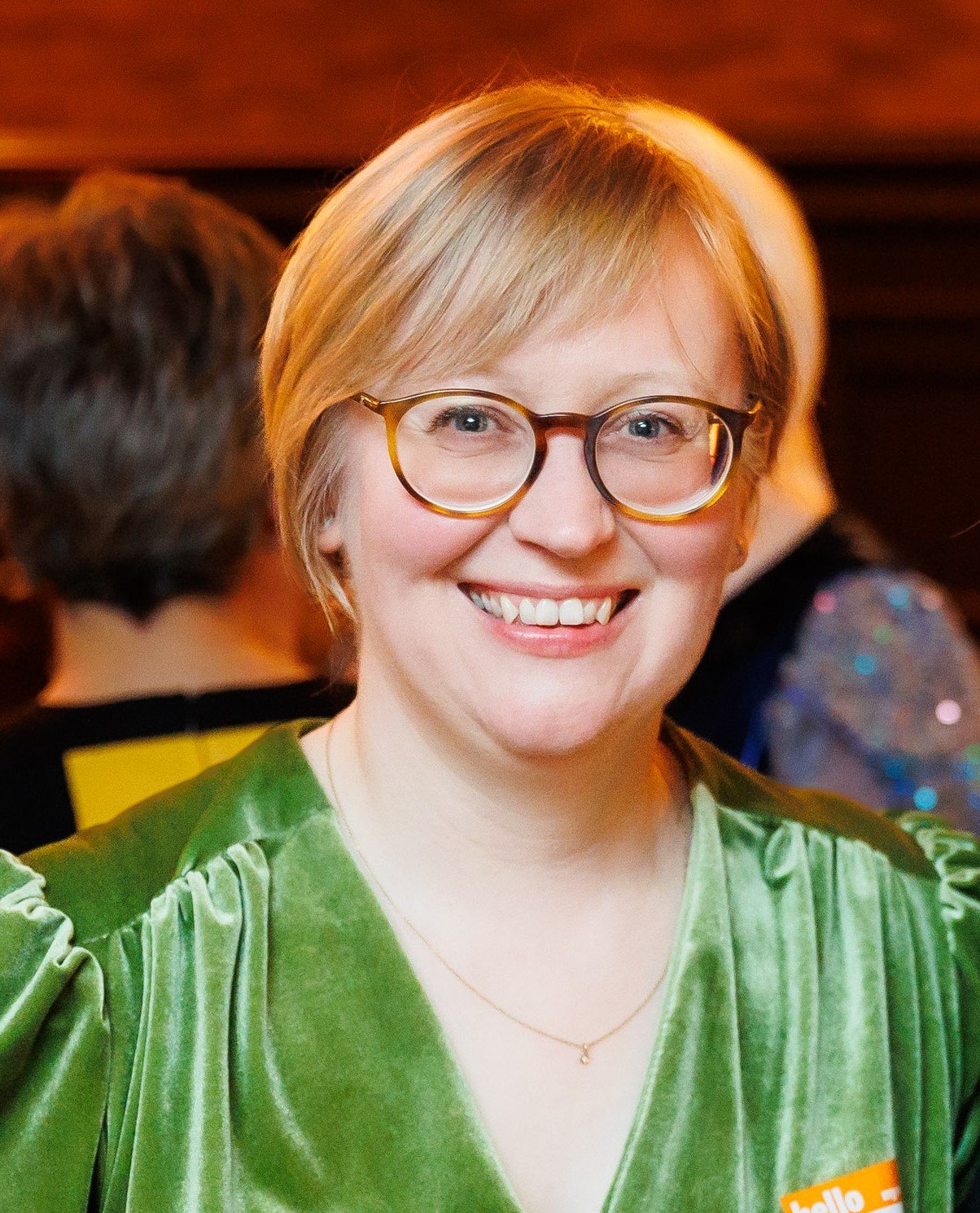 A smiling woman with short blonde hair, wearing glasses with round frames, a green velvet top, and a delicate gold necklace, at a social gathering.