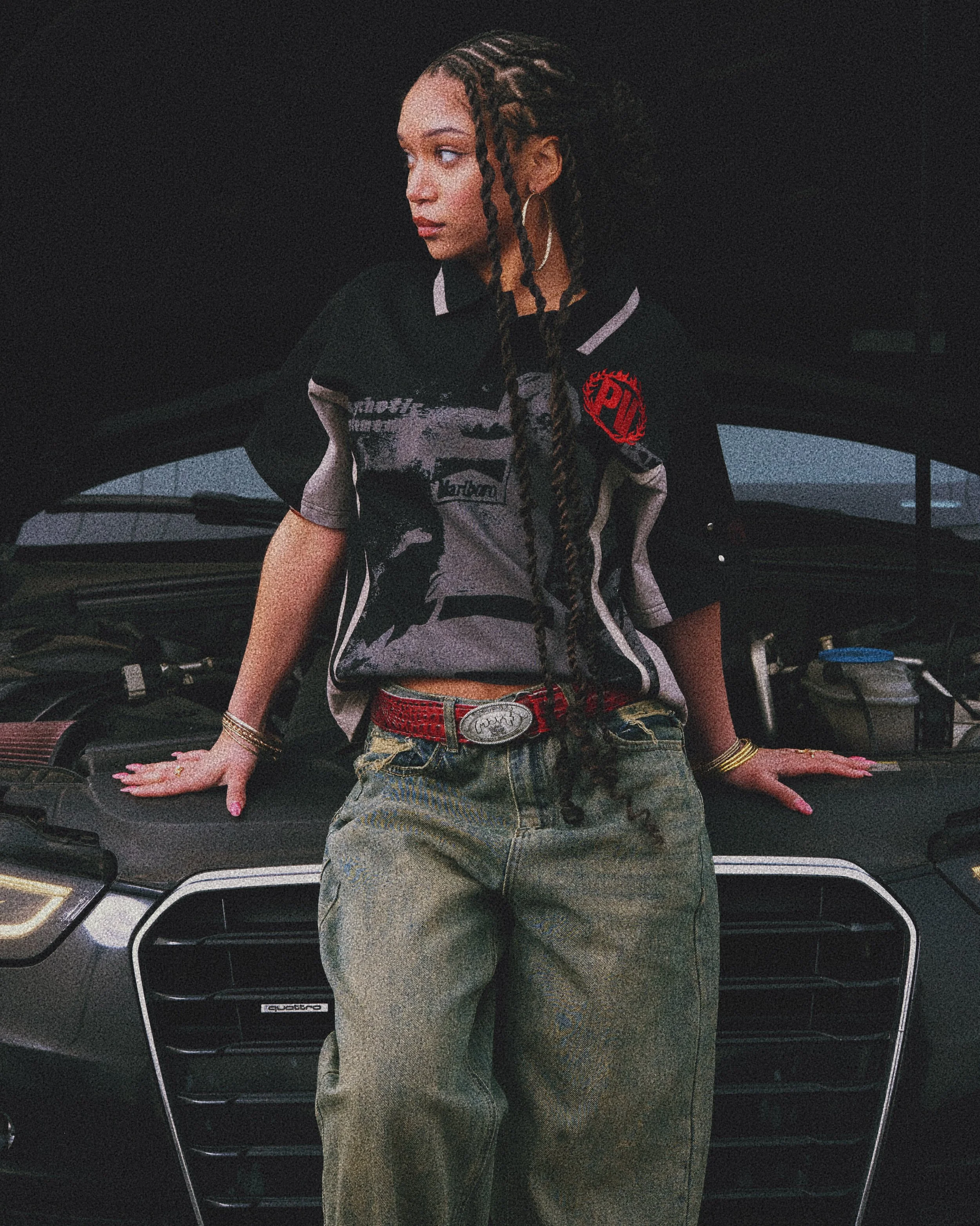 A young woman with long braided hair leaning on the open hood of a black Audi car, wearing a casual graphic polo shirt, baggy jeans, and jewelry.