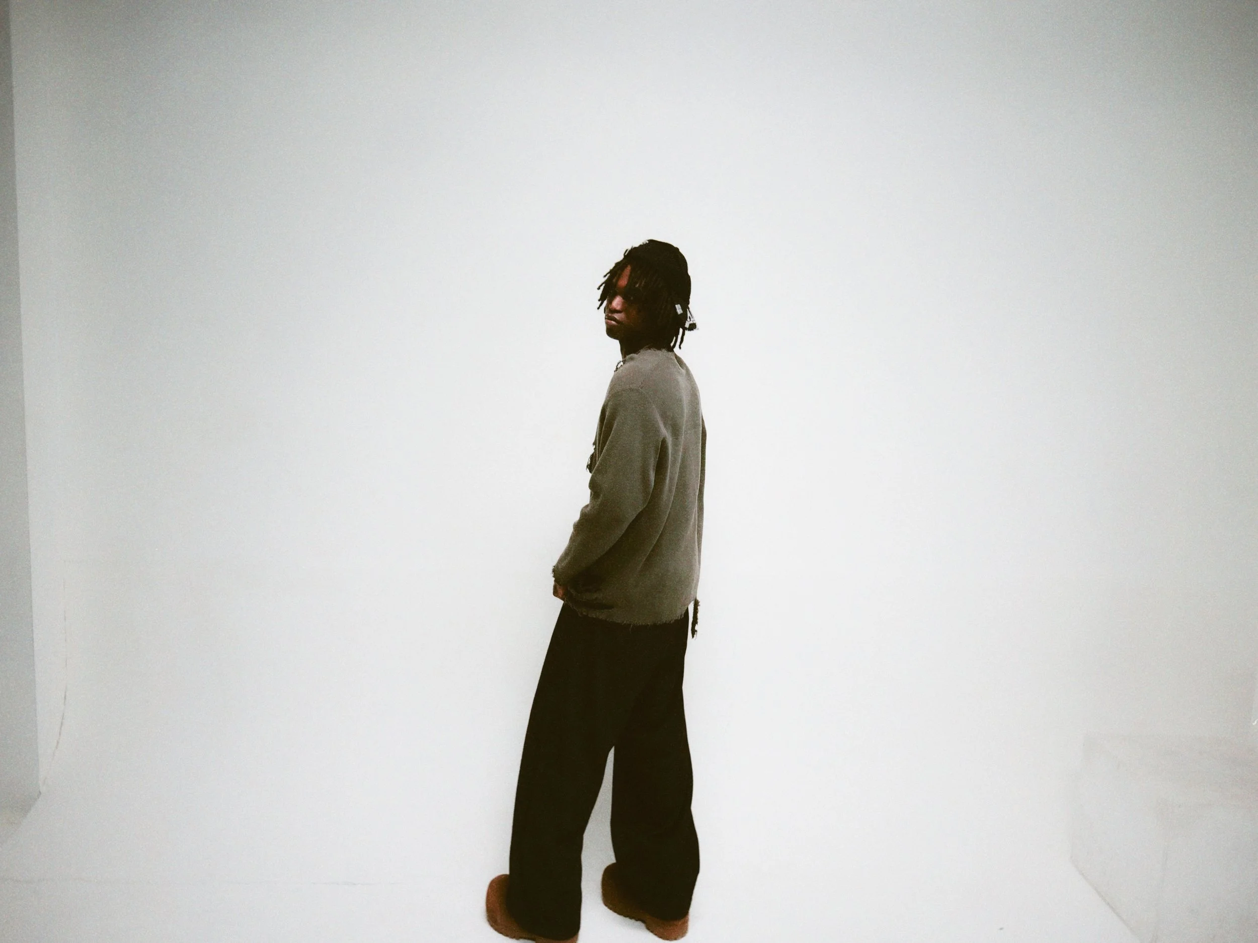 A person with dreadlocks wearing a gray jacket and black pants standing against a plain white wall.
