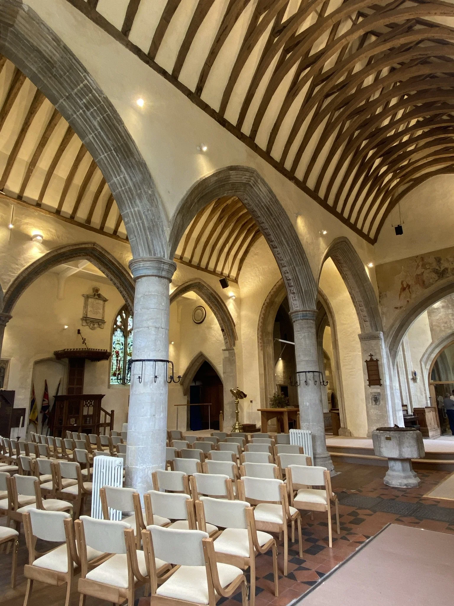 Conservation Architecture — Interior of a medieval church with a wooden vaulted ceiling, stone gothic arches, rows of chairs, stained glass window, and altar area.