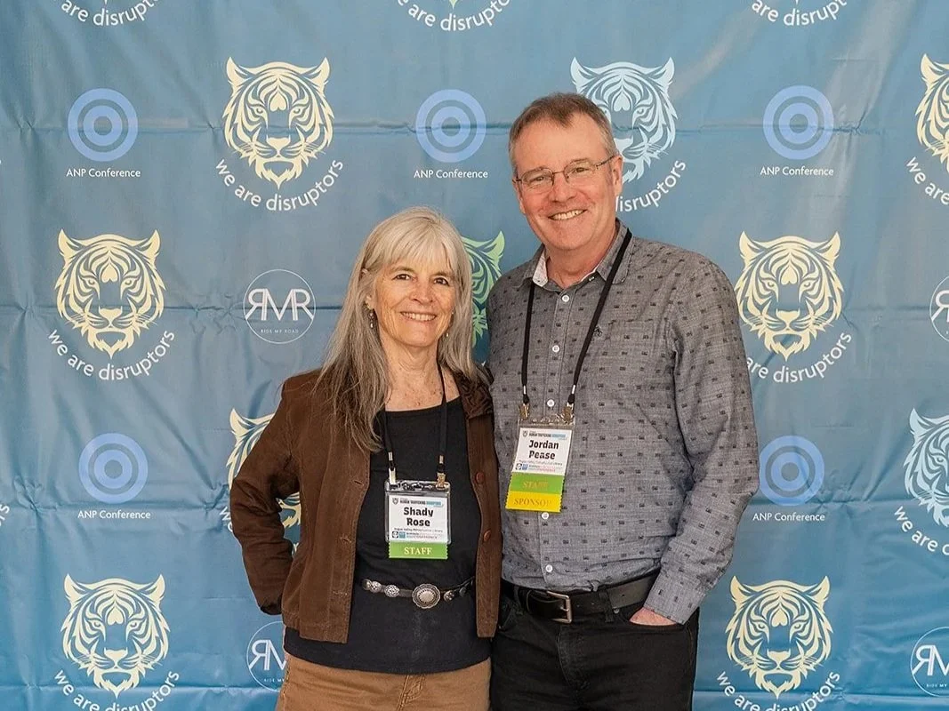 A man and woman standing together at an event in front of a blue backdrop with a tiger logo and the phrases "we are disruptors" and "ANP Conference." The woman has gray hair, wears a brown jacket, black top, beige pants, and black shoes. The man has
