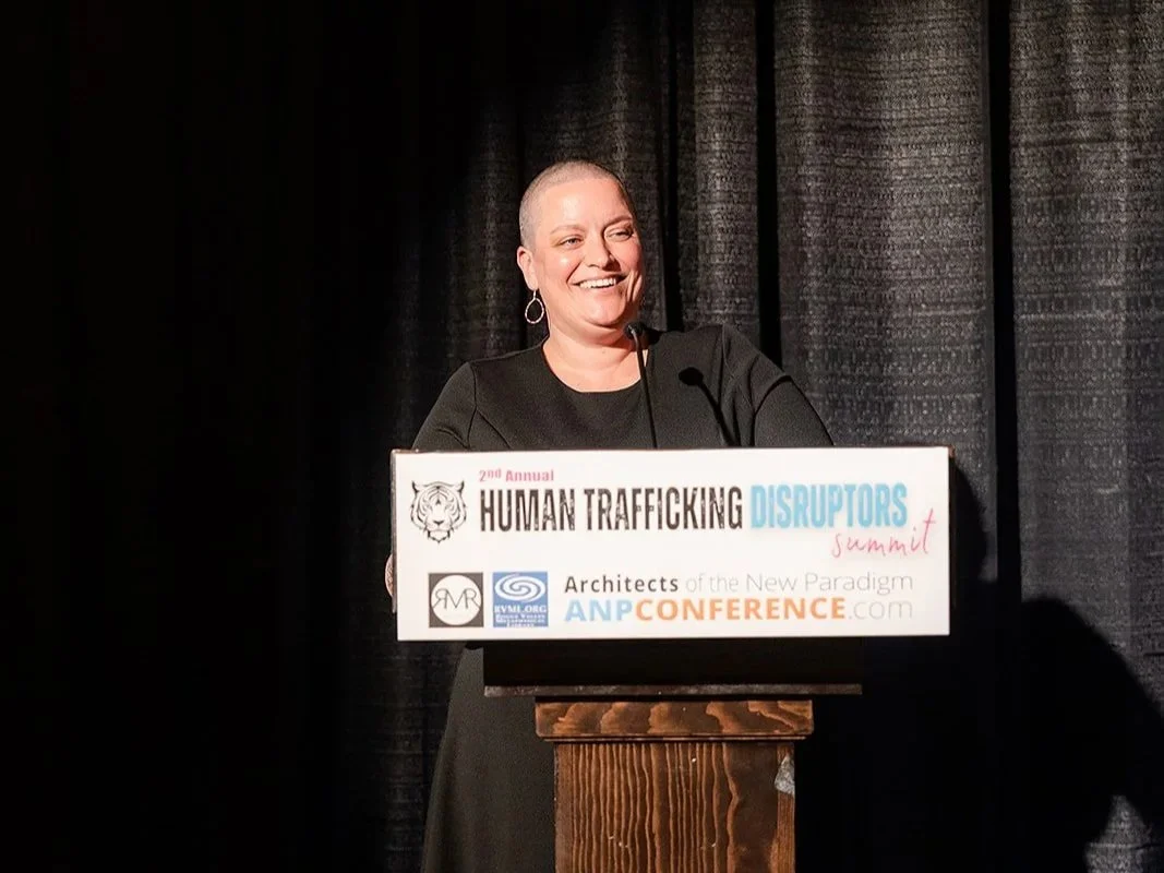 A woman with a shaved head and earrings behind a podium at a conference about human trafficking, with a black curtain in the background.
