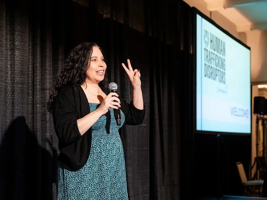 A woman with dark curly hair holding a microphone and making a peace sign while speaking on stage at the National Trapping Dispersers event with a large presentation screen in the background.