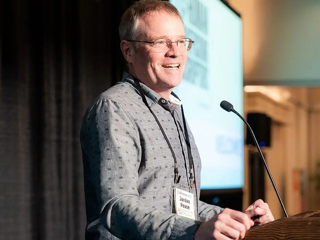 A man with glasses and a gray patterned shirt speaking at a podium during a conference, with a large presentation screen behind him.