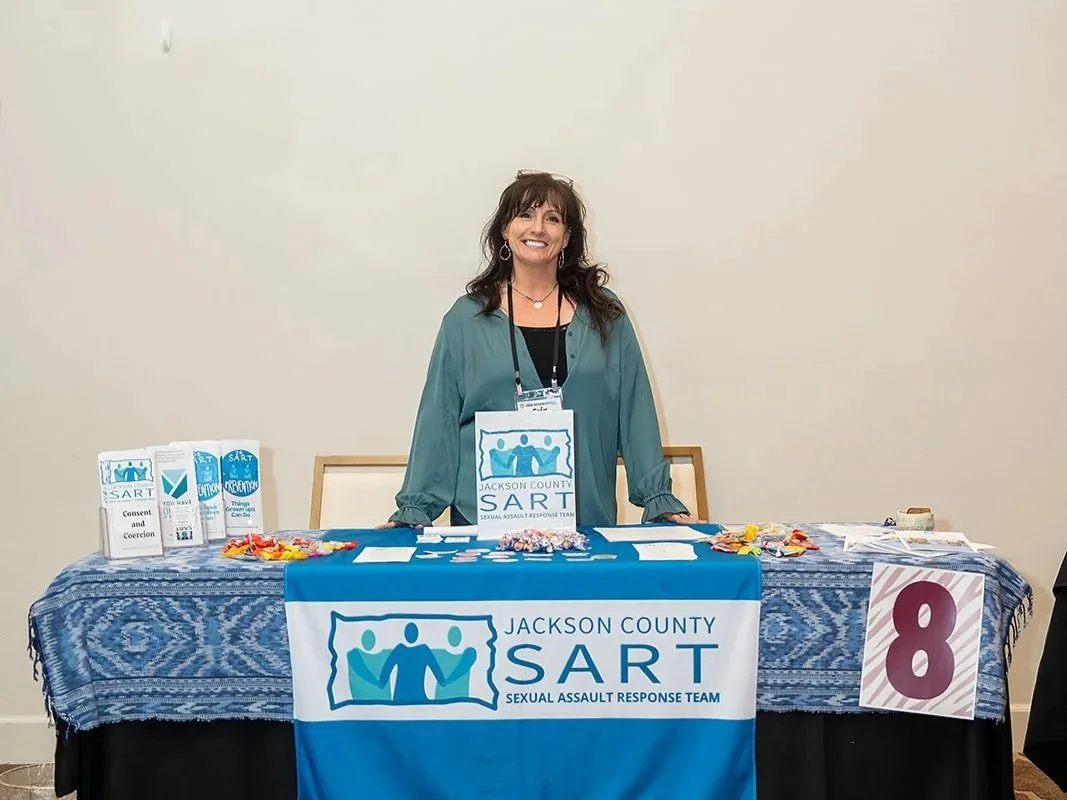 A woman standing behind a table with Jacksonville County SART (Sexual Assault Response Team) banner at a conference or event, with informational brochures, candies, and a sign showing the number 8 on the table.