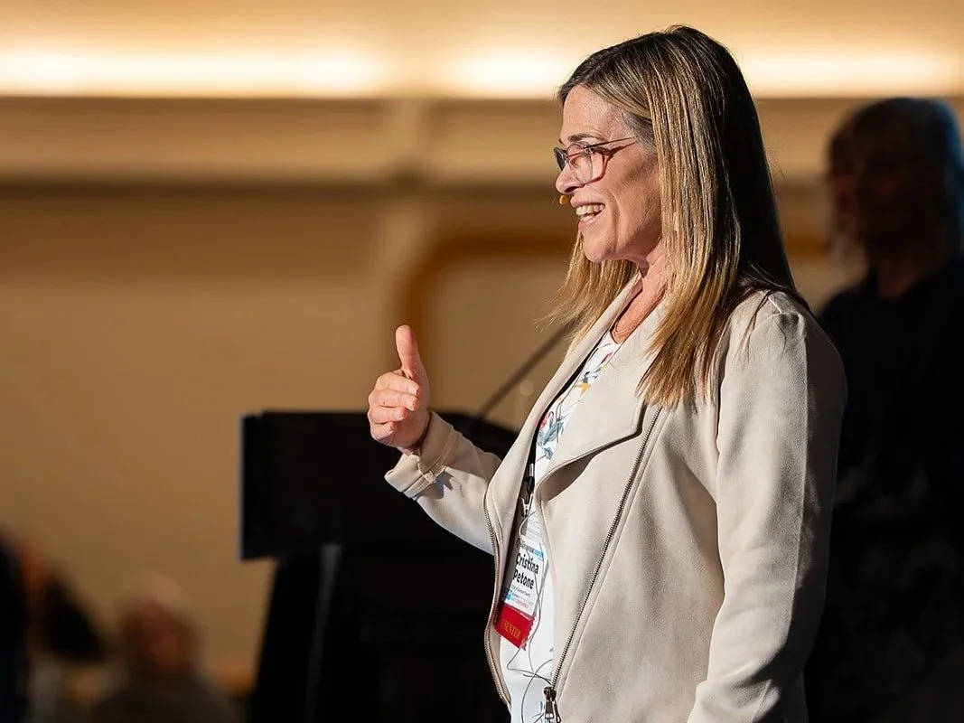 A woman speaking at a conference, holding green folders, giving a thumbs-up, wearing glasses, a beige jacket, and red pants, with a microphone headset.