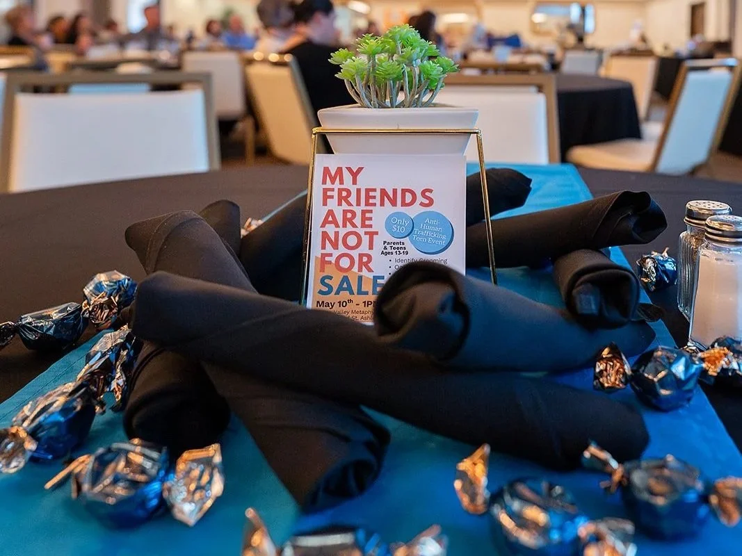 A table centerpiece in a banquet hall with a small potted plant, an informational sign about a campaign against human trafficking, wrapped candies, and black napkins on a blue tablecloth, with other tables and guests in the background.