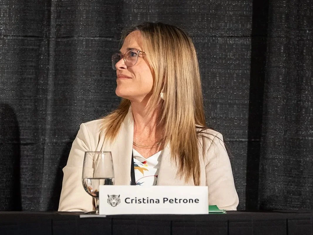 A woman with blonde hair wearing glasses and a cream blazer sitting at a table with a name tag that reads 'Cristina Petrone', a glass of water, and a black tablecloth, against a black curtain backdrop.