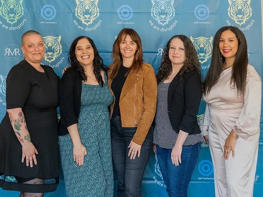 Five women standing in front of a blue backdrop with tiger and logo graphics, at an event called ANP Conference, smiling at the camera.