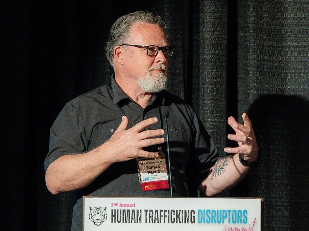A man with glasses and a beard speaking at a podium during a conference about human trafficking disruptors. The podium has a sign that reads '2nd Annual Human Trafficking Disruptors Summit' and 'Architects of the New Paradigm ANP Conference.' The man