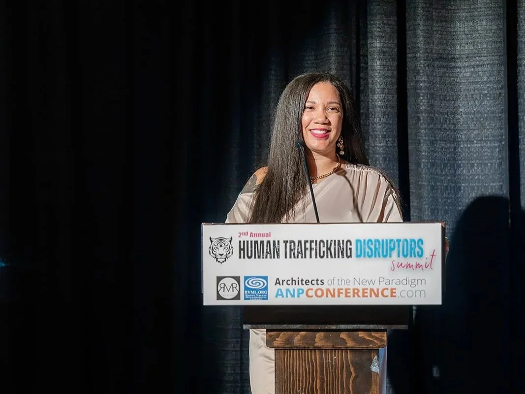 A woman with long dark hair is smiling while speaking at a podium during the 2nd Annual Human Trafficking Disruptors Summit. The podium has a sign with event details, and she is standing in front of dark curtains.