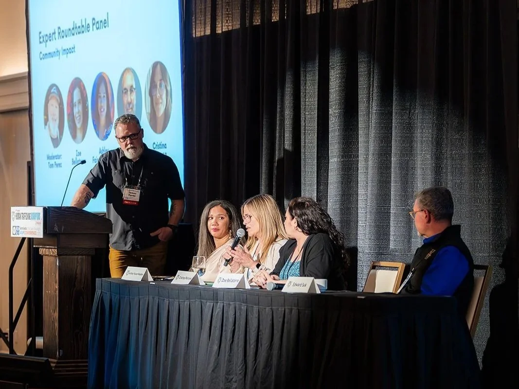 A man standing at a podium speaking to an audience with four women seated at a long table on stage, and a large screen displaying a presentation slide behind them at a conference or panel discussion.