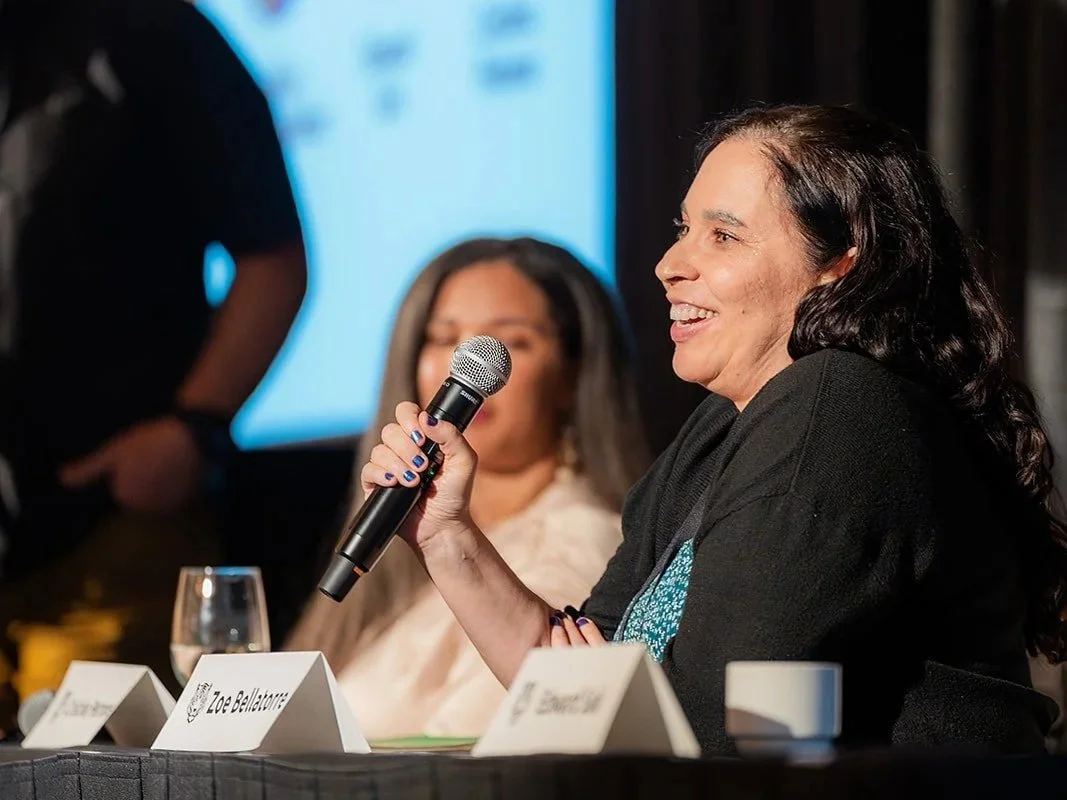 A woman speaking into a microphone at a panel discussion, with a woman in the background and a presentation screen showing images of food.