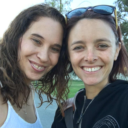 Two women smiling outdoors, one with sunglasses and a black hoodie, the other with a white shirt, in a park or open area.
