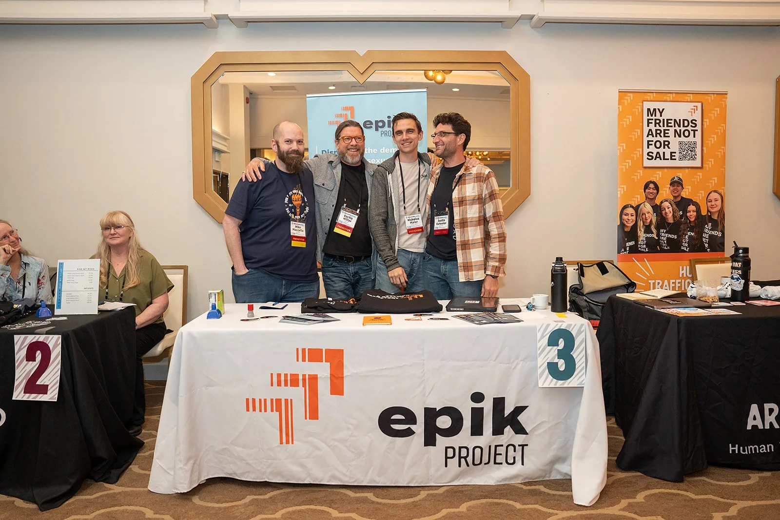 Four men standing together behind a table displaying the Epik Project logo, all smiling at a conference or event.