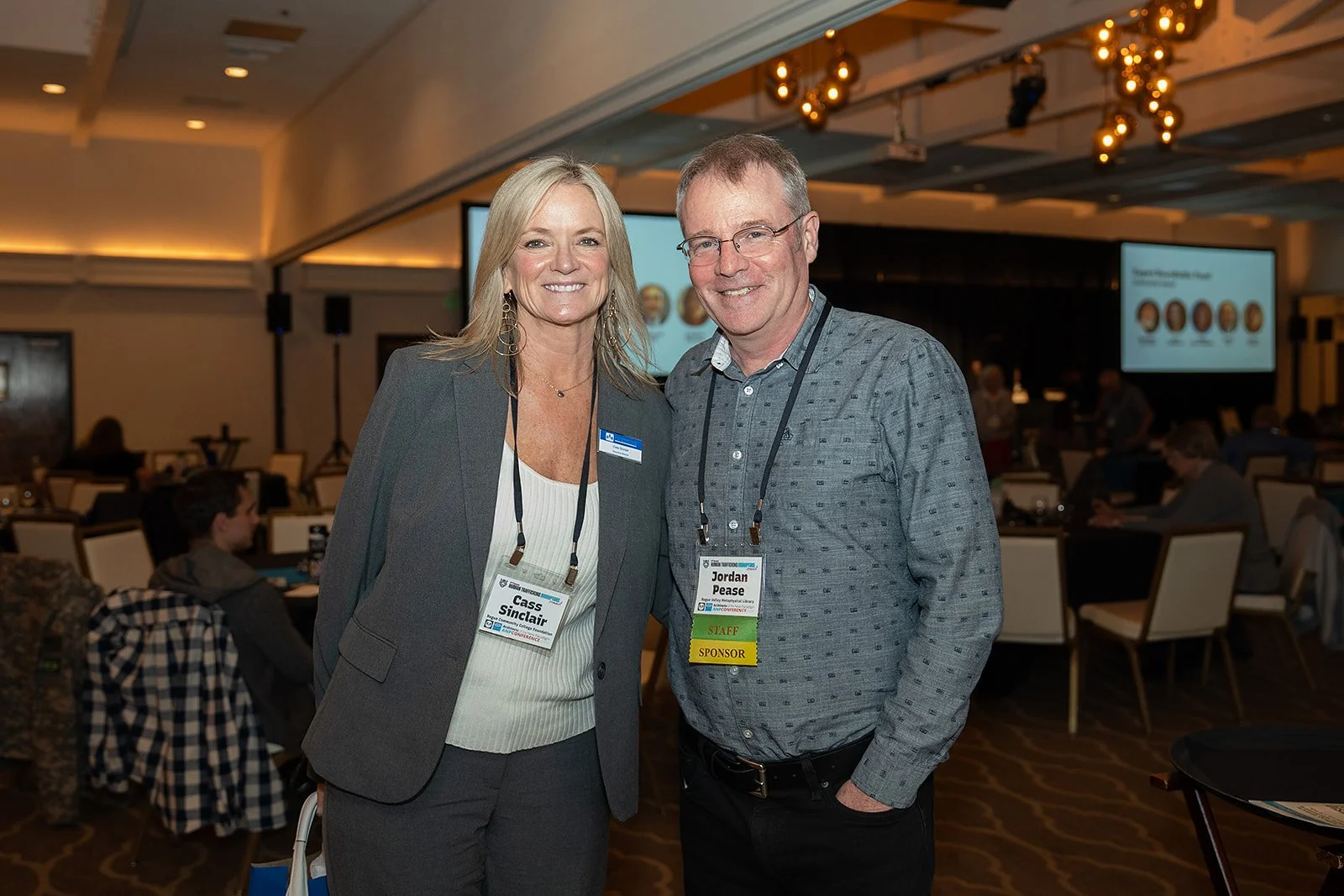 A woman and a man smiling at the camera at a conference. The woman wears a gray blazer and the man a gray shirt. Both have name tags around their necks.