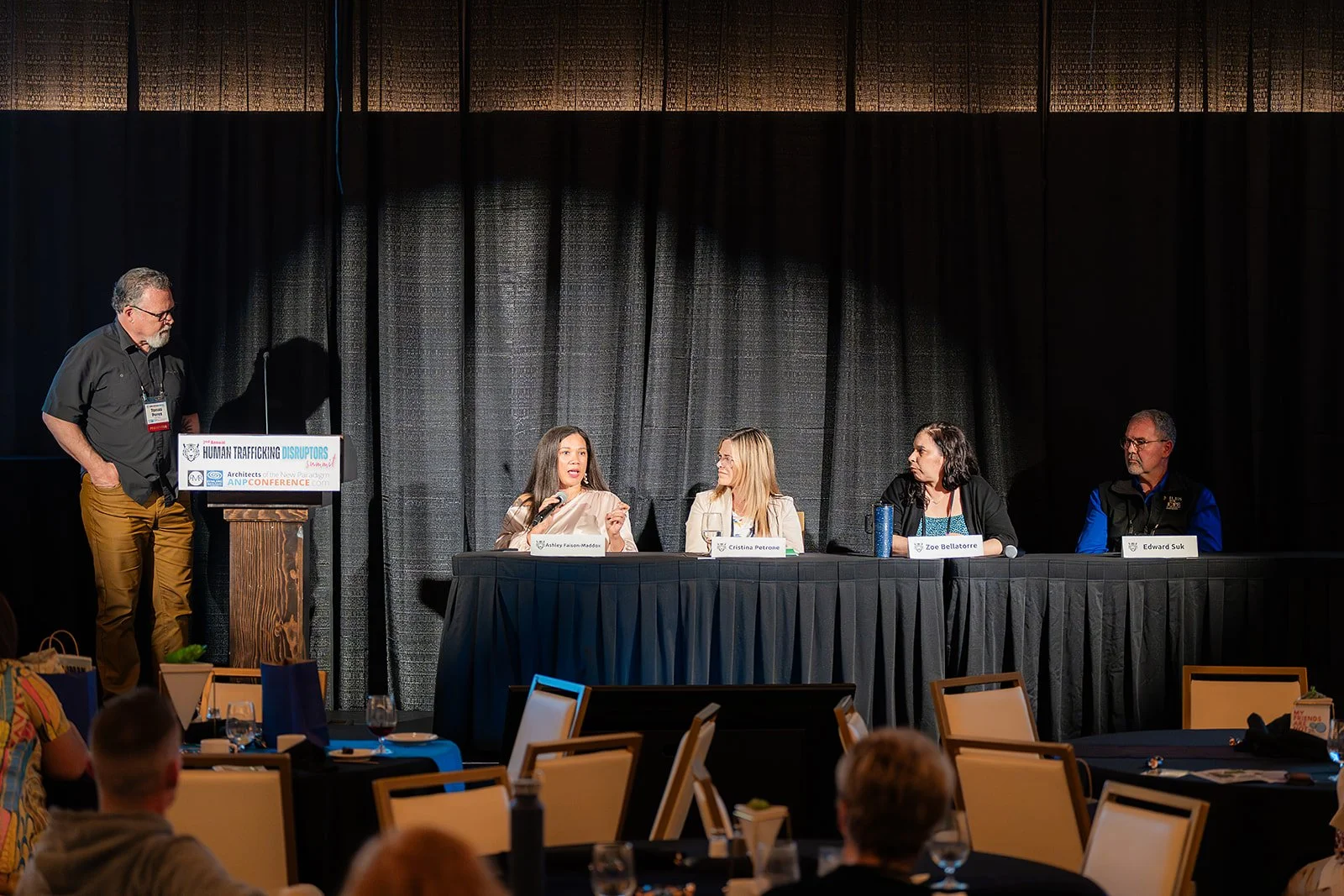 Panel discussion at a conference with five people, four women and one man, on stage, with a black curtain backdrop and an audience in front. A man stands at a podium with a sign about human trafficking and disruptors.