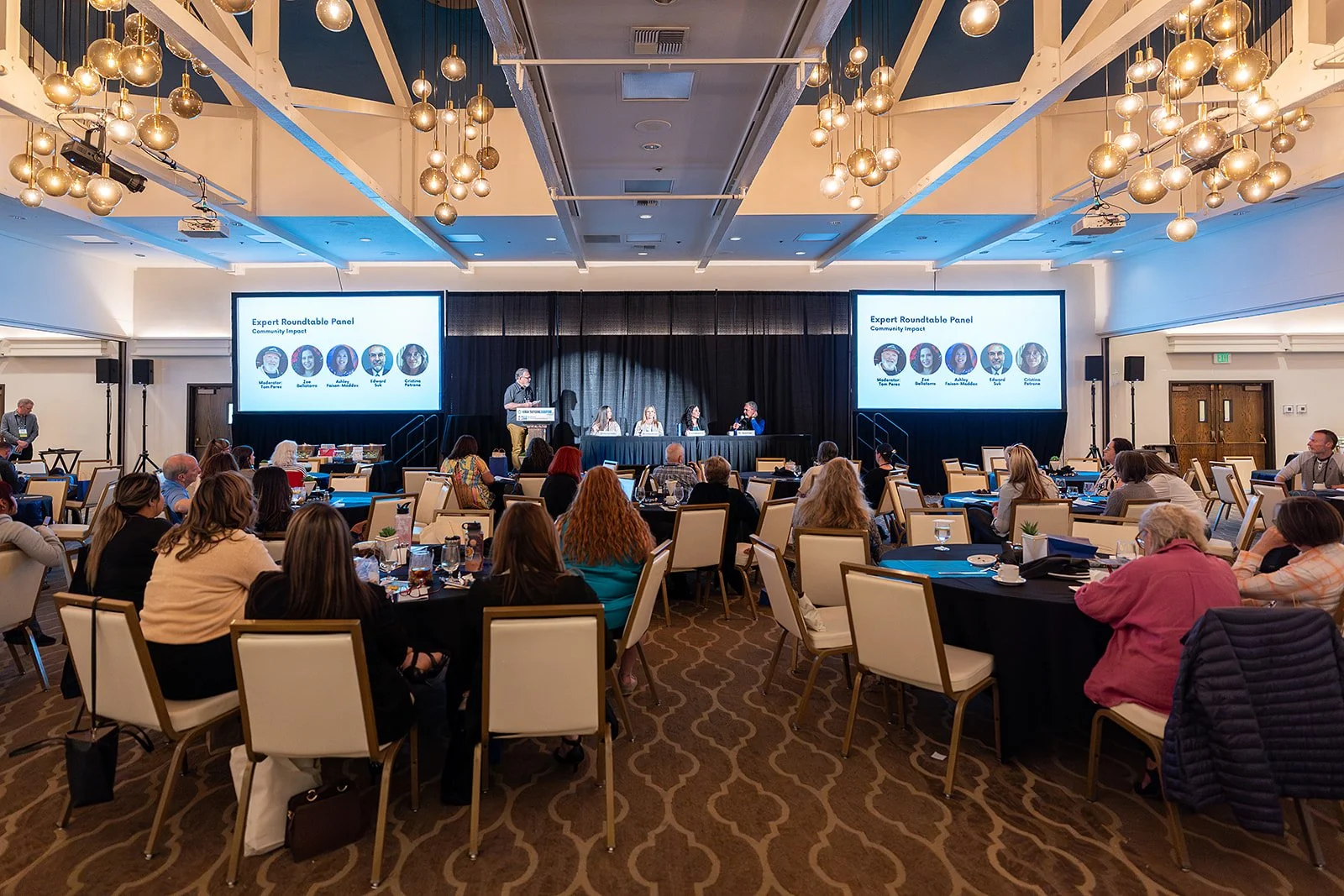 A conference room filled with seated attendees listening to a panel discussion on a stage. The stage has a podium and five panelists seated at a long table, with large screens on either side displaying the panel members' names and photos. The ceiling