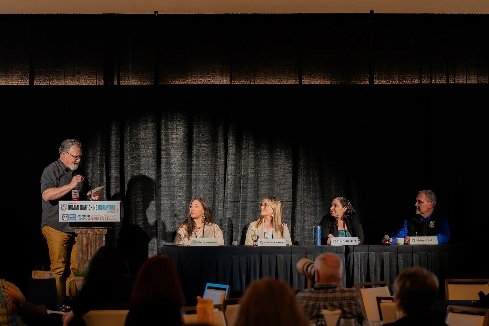 A panel discussion at a conference with five people, one man at a podium on the left and four women and men seated at a long table on the right, in front of a black curtain background.