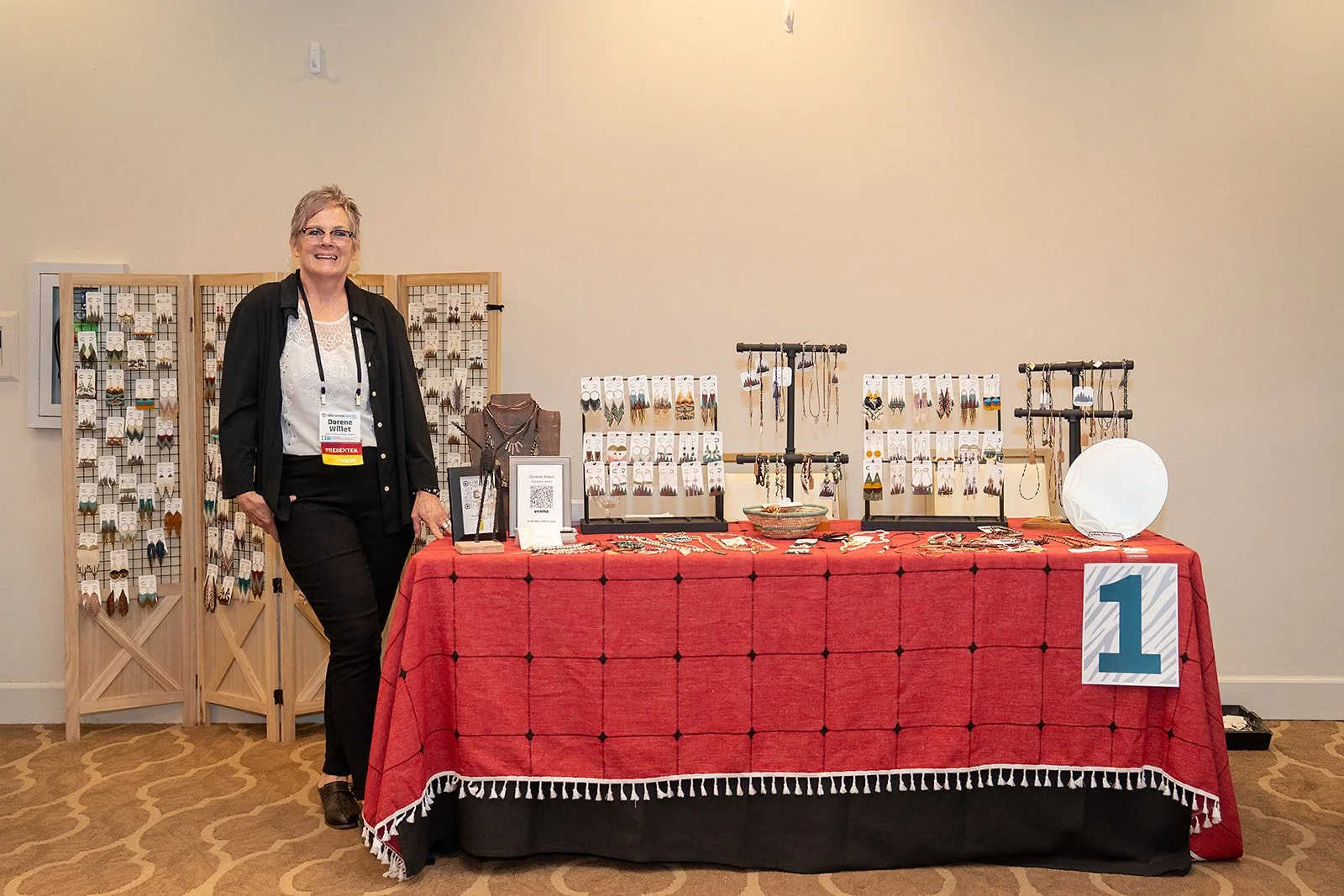 A woman wearing glasses and a black jacket stands next to a display table with jewelry at a craft fair or exhibition. The table has a red tablecloth and is set up with various earrings, necklaces, and bracelets, with another display of earrings on bl