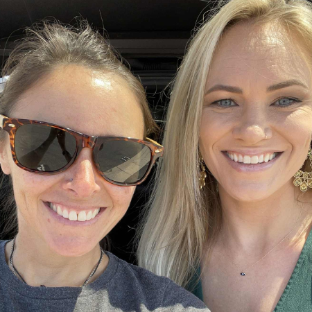 Two women smiling for a selfie, one wearing sunglasses and the other wearing earrings.