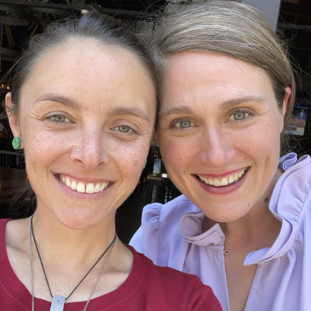 Two women smiling closely for a selfie, one with dark hair and green earrings, the other with light hair and a lavender blouse.