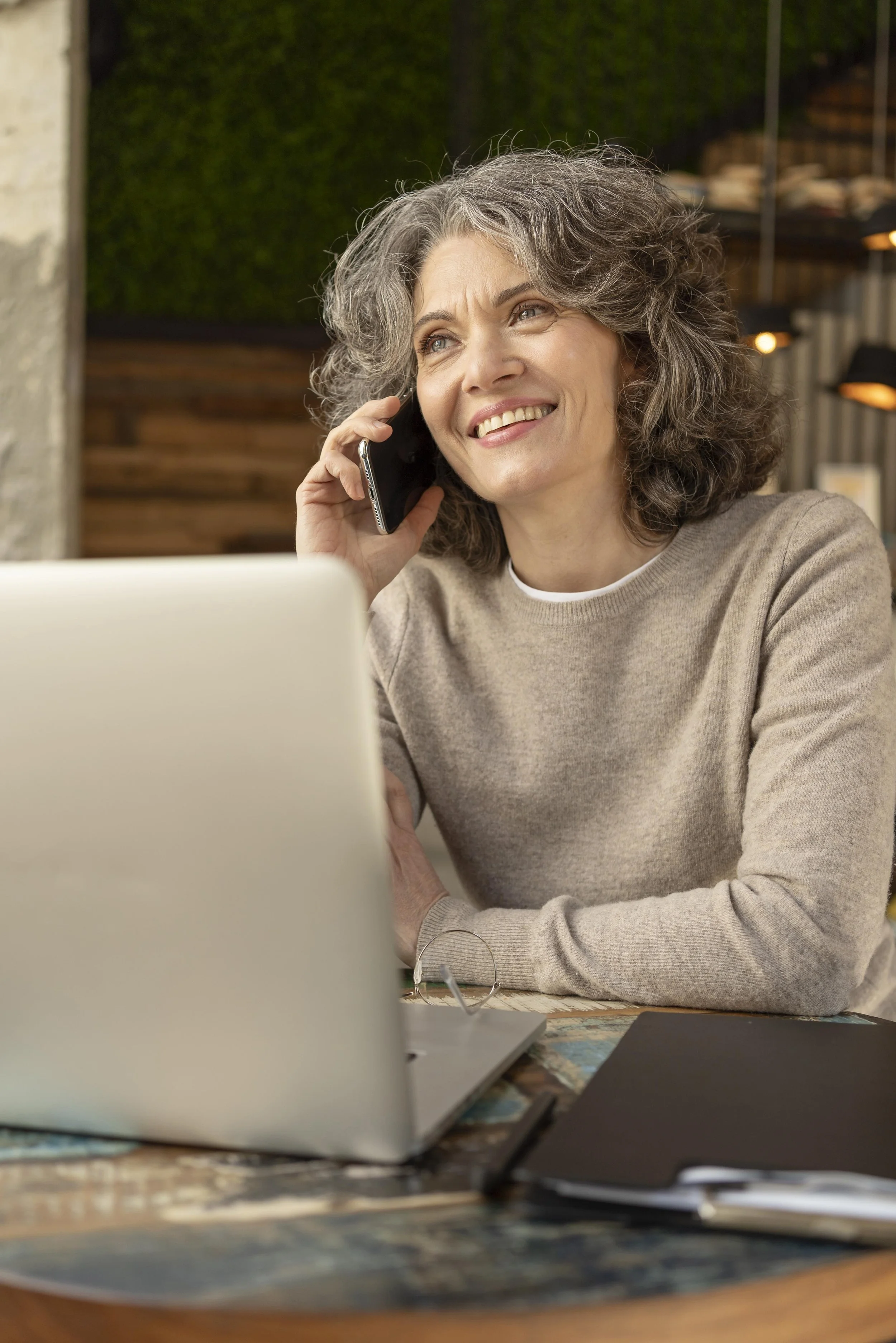 A woman with curly gray hair smiling and talking to Karen her fearless Journey life coach on her cellphone while sitting at a desk with a laptop and notebooks talking about the online course she just completed.