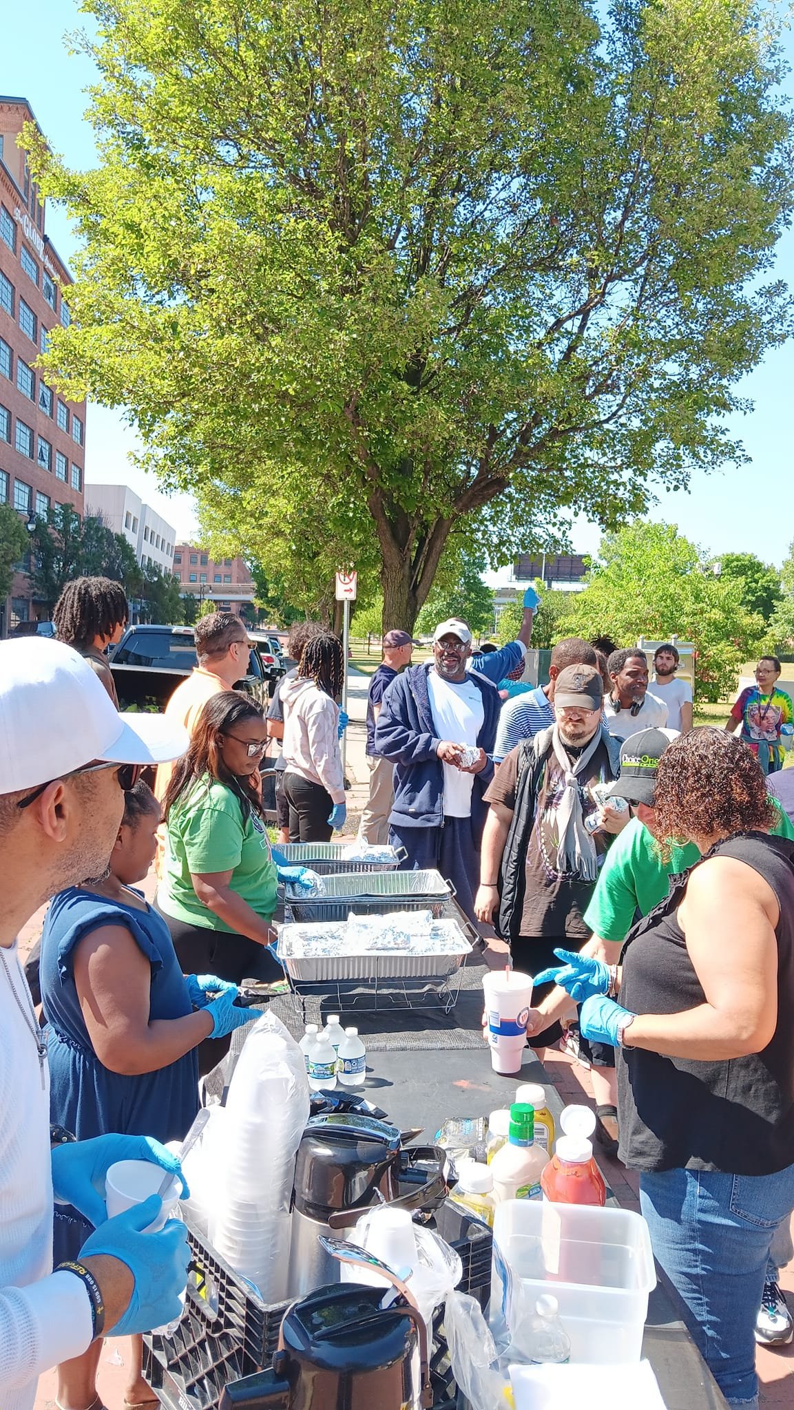 Group of people gathered outdoors around a table with food and supplies, with a large tree and buildings in the background.