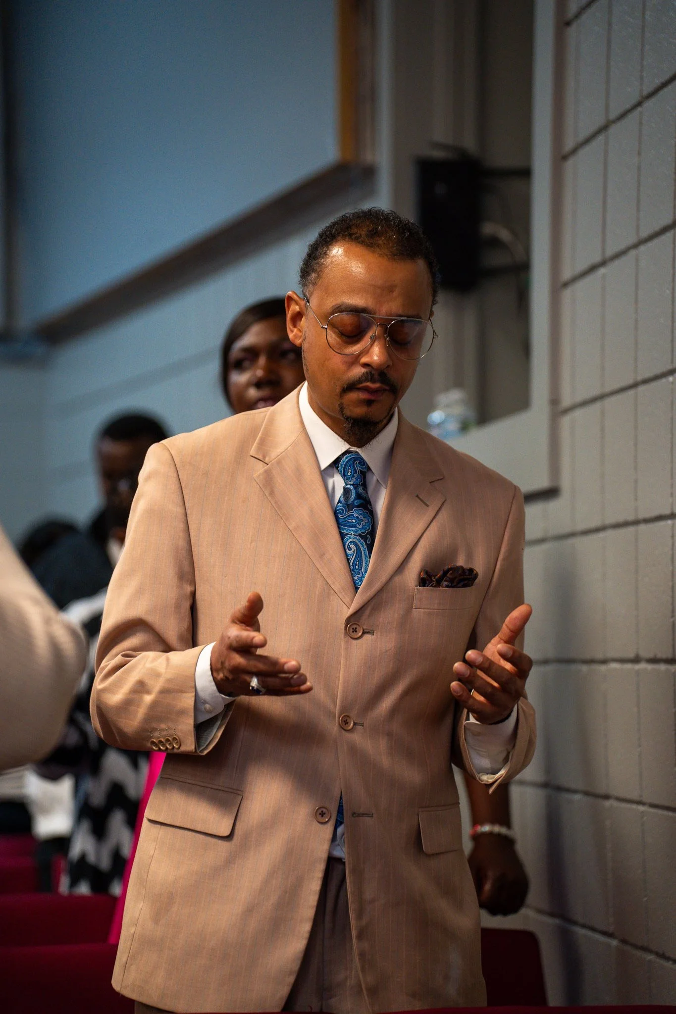 Man in a beige suit with glasses and a blue patterned tie praying in a church.
