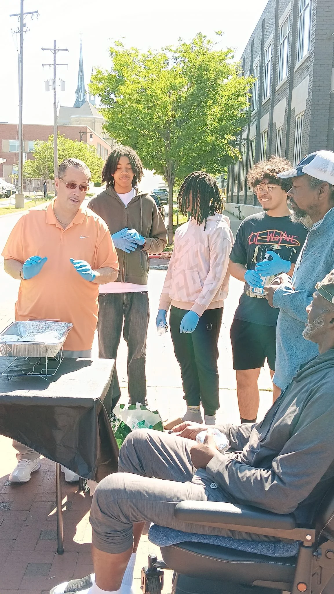 A group of people gathered outdoors near a tree and a building, engaged in a discussion or activity, with some wearing blue gloves and a table with food or supplies in front of them.