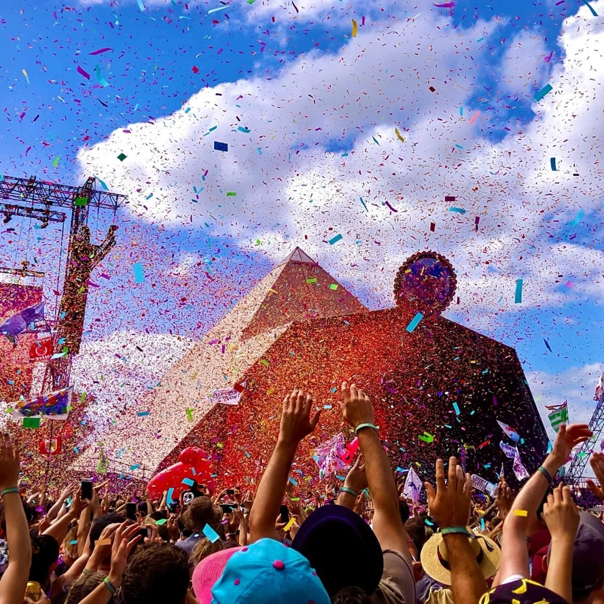 Crowd celebrating with colorful confetti and streamers at an outdoor festival, with a large pyramid-shaped structure and a Ferris wheel in the background.