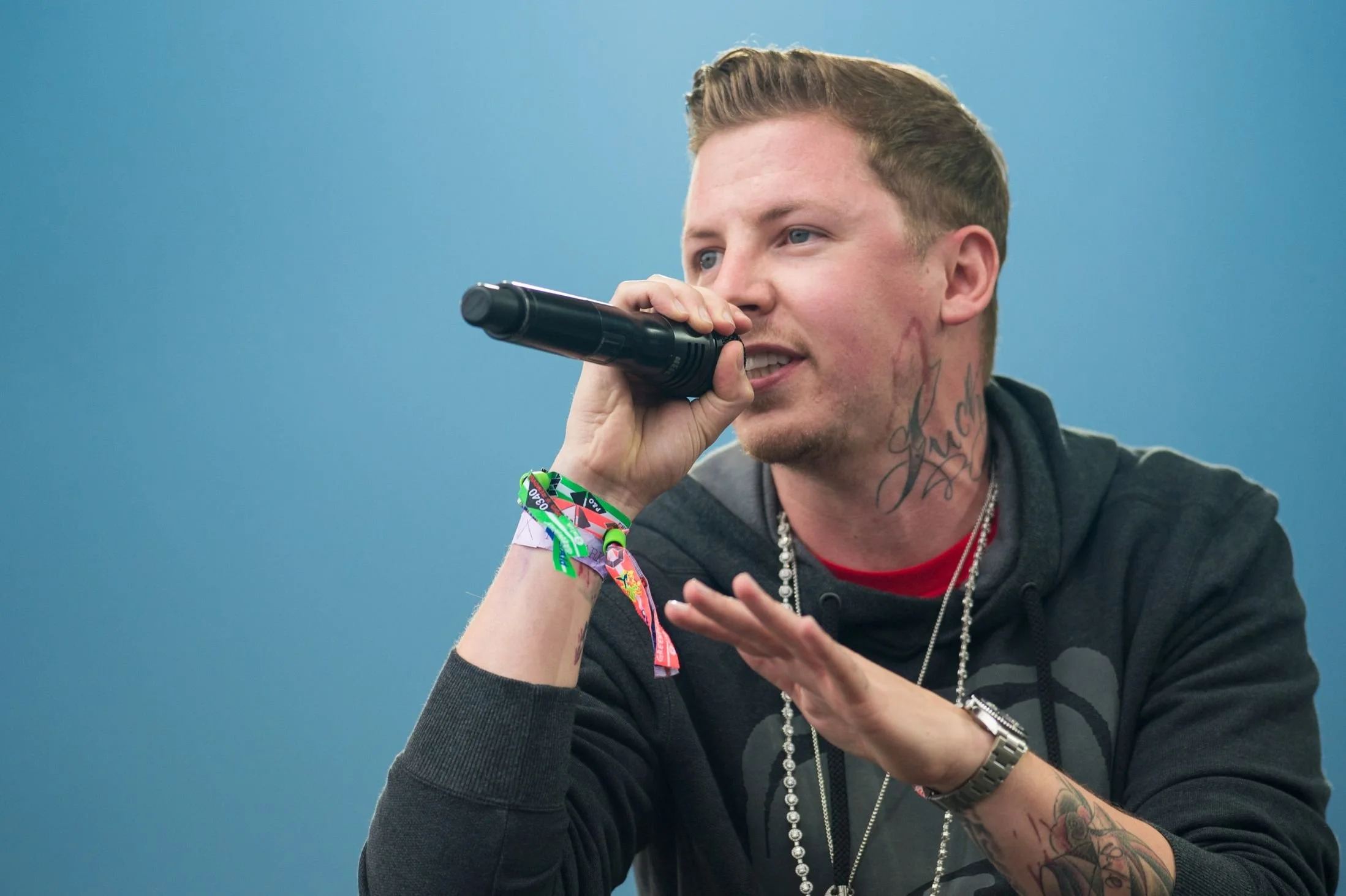 A man with tattoos, wearing a dark hoodie and jewelry, speaking into a microphone against a blue background.