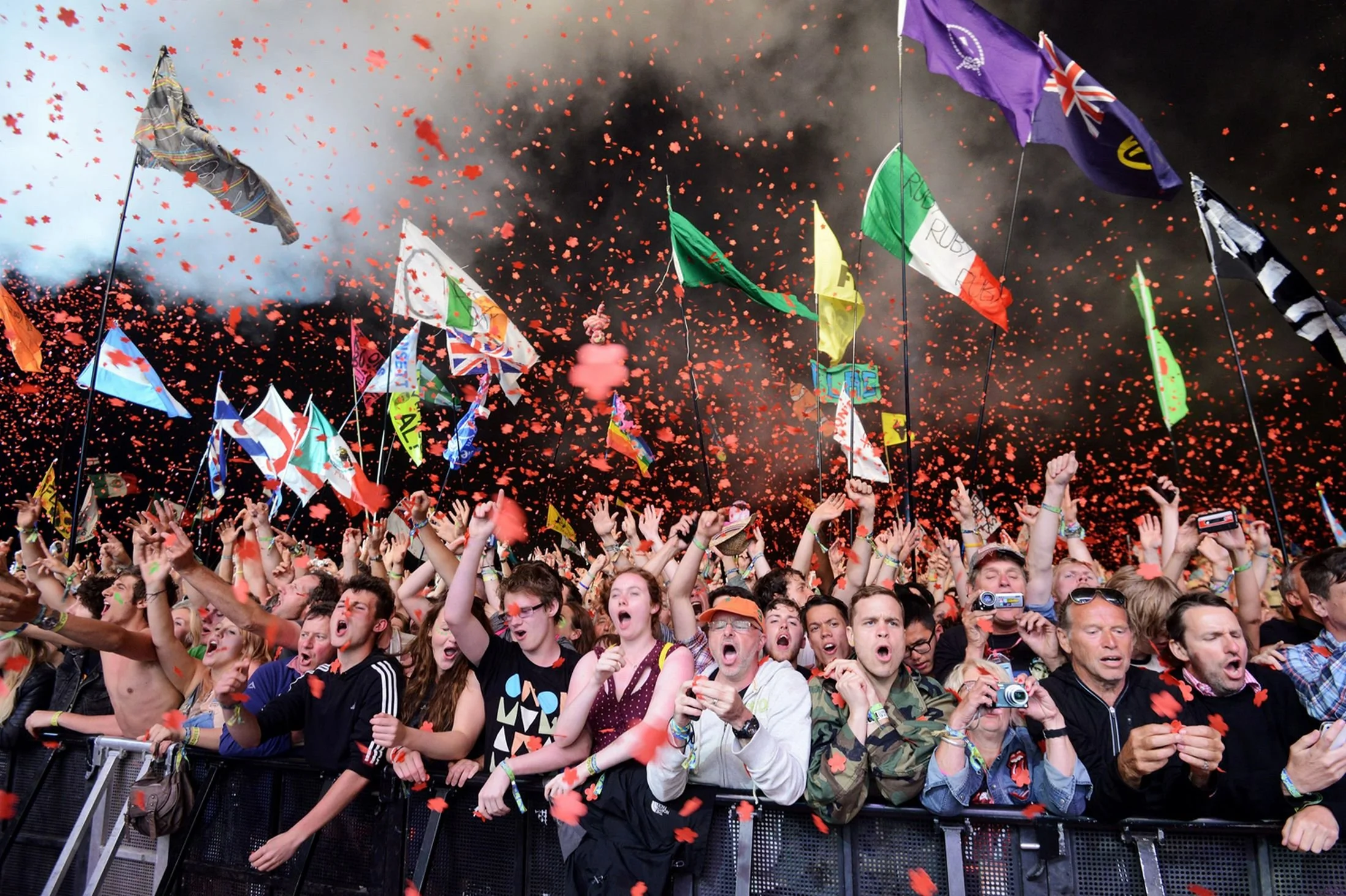 Crowd of people celebrating at a concert or festival, with many holding flags and waving, red confetti falling from above, and a dark background.