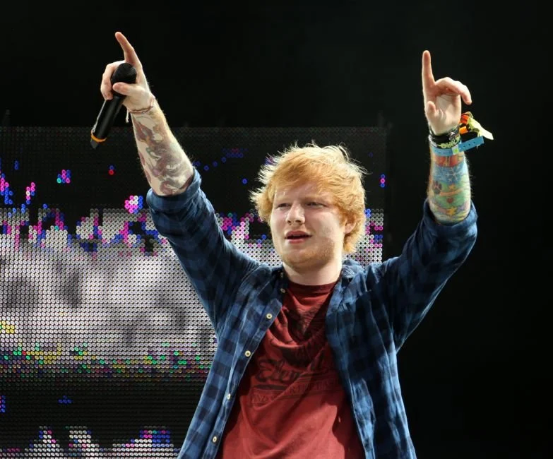 A young man with blonde hair and tattoos is on stage holding a microphone in his right hand, raising his left hand with his index finger pointing up, with a colorful LED backdrop behind him.