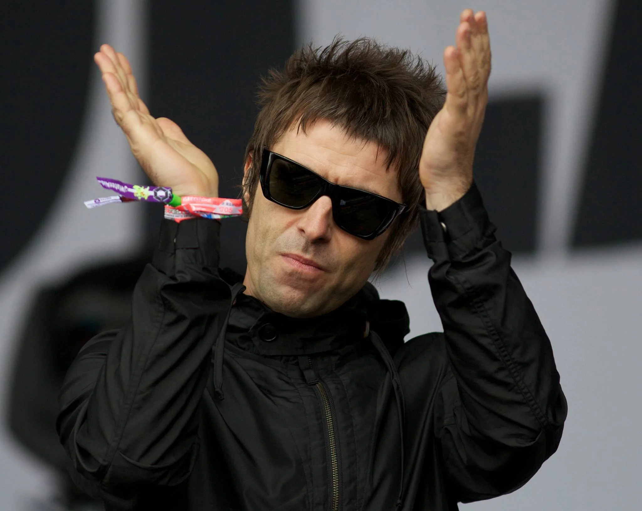 A man wearing black sunglasses and a black jacket, making a gesture with hands raised and palms open, with wristbands on his wrist.