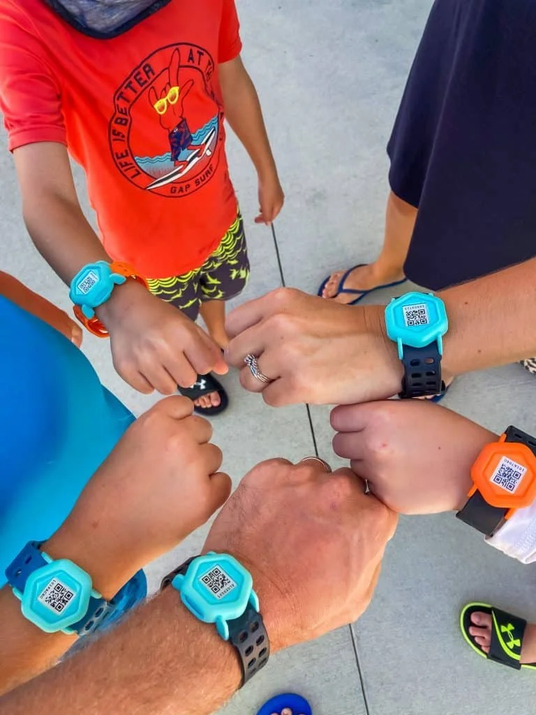 Group of people showing their colorful smartwatches with QR codes, with a boy in a red graphic T-shirt standing among them.