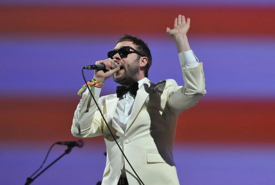 Male performer wearing sunglasses and a cream-colored tuxedo jacket with a bow tie, singing into a microphone with his right hand raised.