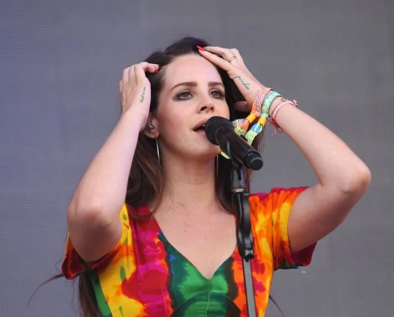A woman with long dark hair singing into a microphone, wearing a colorful tie-dye shirt and various bracelets, with a neutral background.