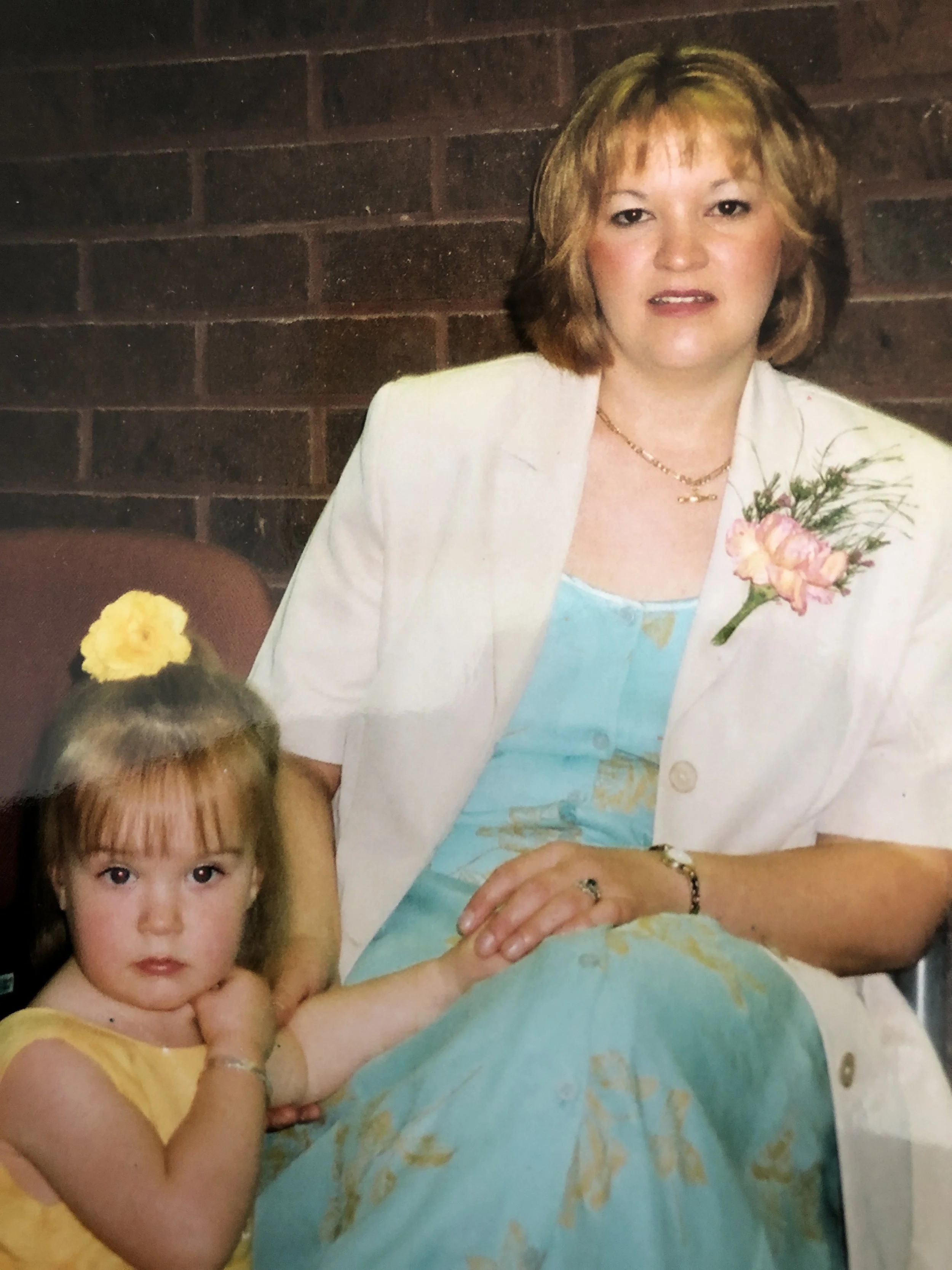 A woman with short, light brown hair sitting next to a young girl with long brown hair, wearing a yellow dress. The woman is in a white blazer with a pink flower corsage, sitting against a brick wall. The girl is looking at the camera and is holding the woman's hand.