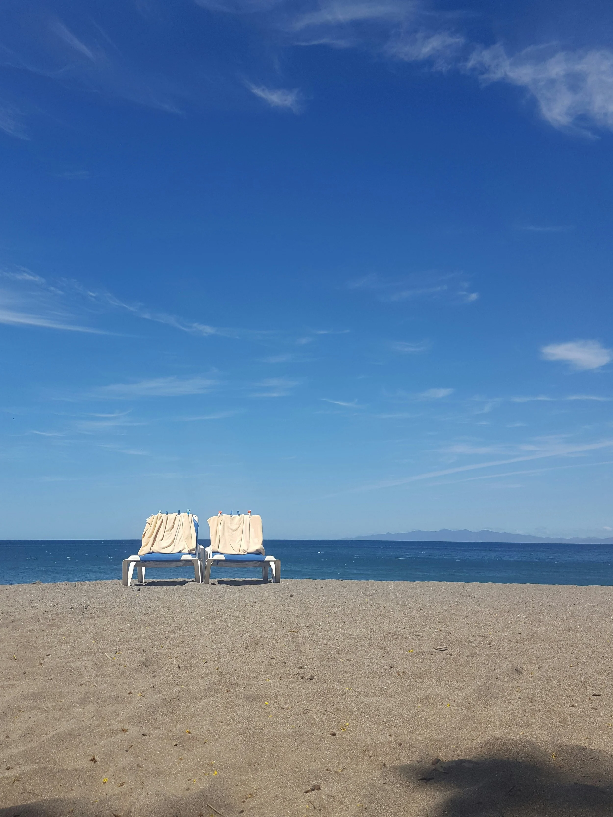 Two empty beach chairs covered with towels on sandy beach facing the ocean under a blue sky with some clouds.