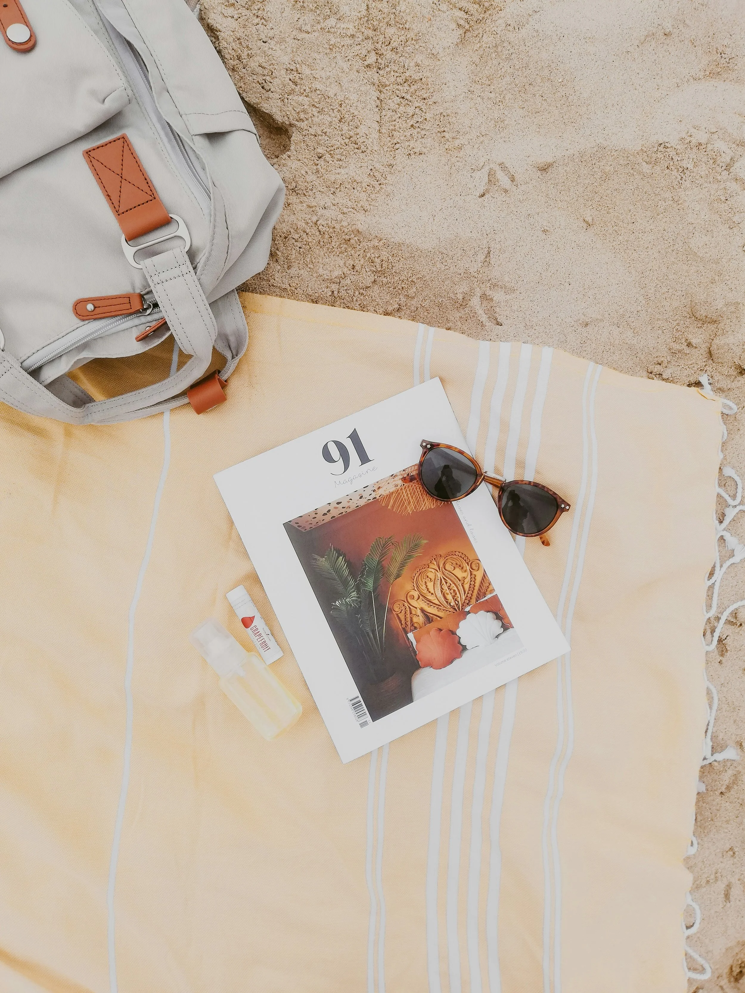 Beach scene with a beige striped towel on the sand. On the towel, there is a magazine, a pair of sunglasses, a small tube of sunscreen, and a white backpack with brown accents.