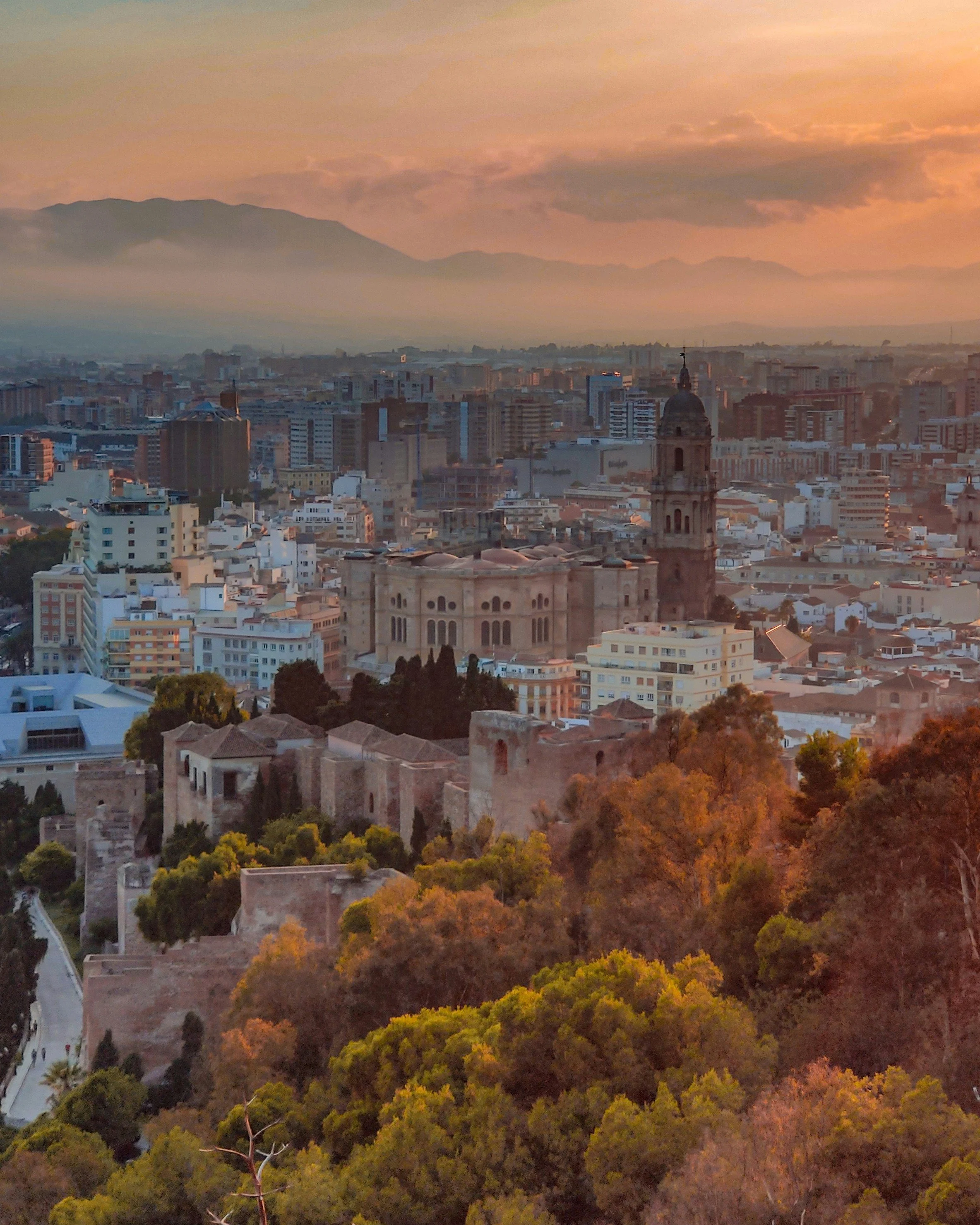 Aerial view of a city at sunset with mountains in the background, featuring historic and modern buildings, including a church with a tall bell tower, surrounded by trees with autumn foliage.
