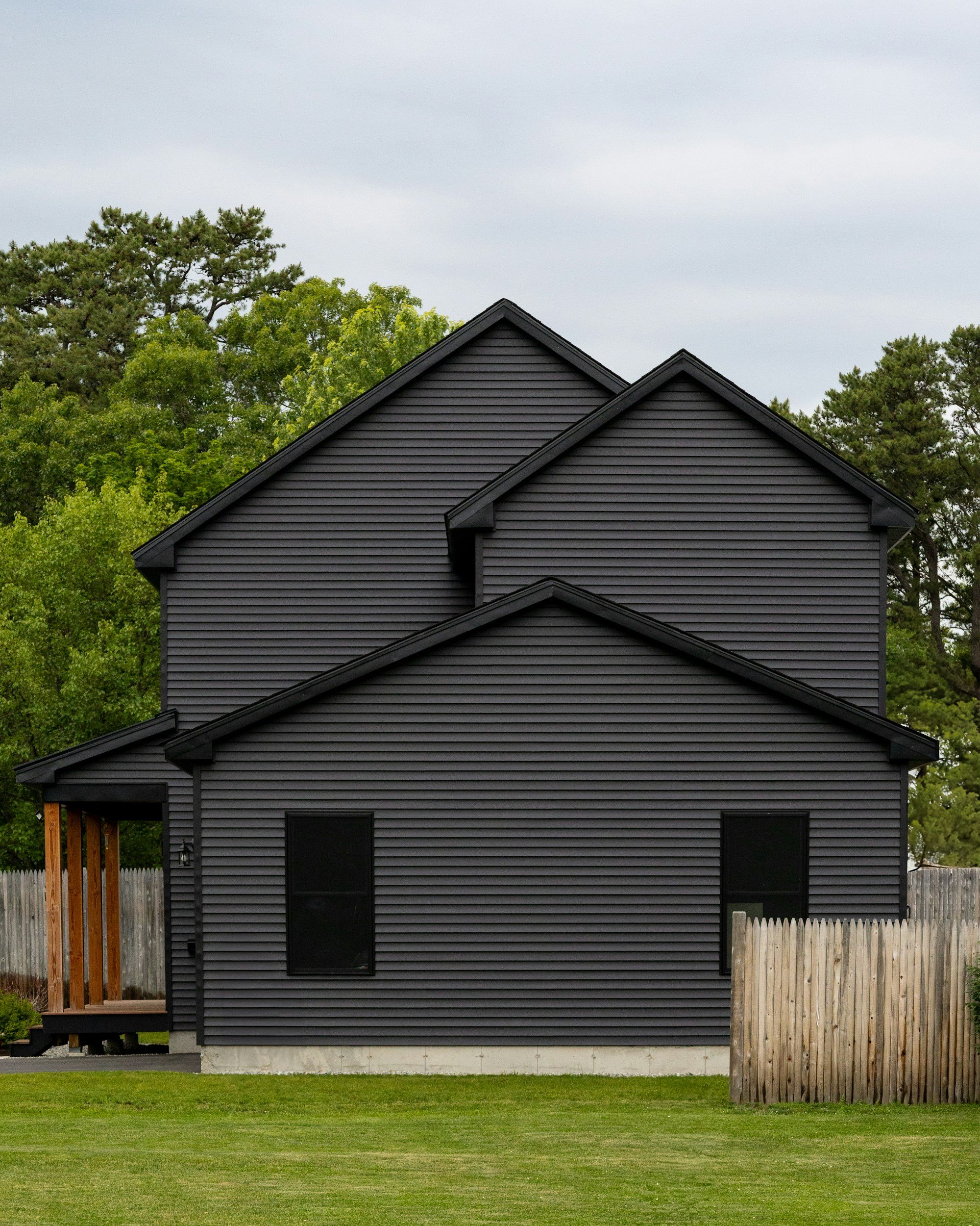 A black farmhouse-style house with black vinyl siding, a small porch with wooden posts, and a wooden fence, surrounded by green grass and trees under a cloudy sky. DentonPaint.Com is your go to painter in Denton County and surrounding areas.