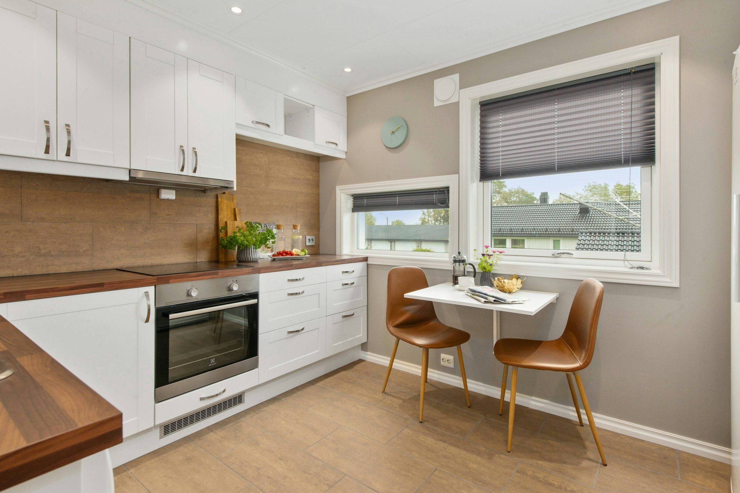 Kitchen with white cabinets, wooden countertops, two large windows with dark blinds, two brown chairs around a small table, and some plants and decorations on the counter. DentonPaint.Com is your go to painter in Denton County and surrounding areas.