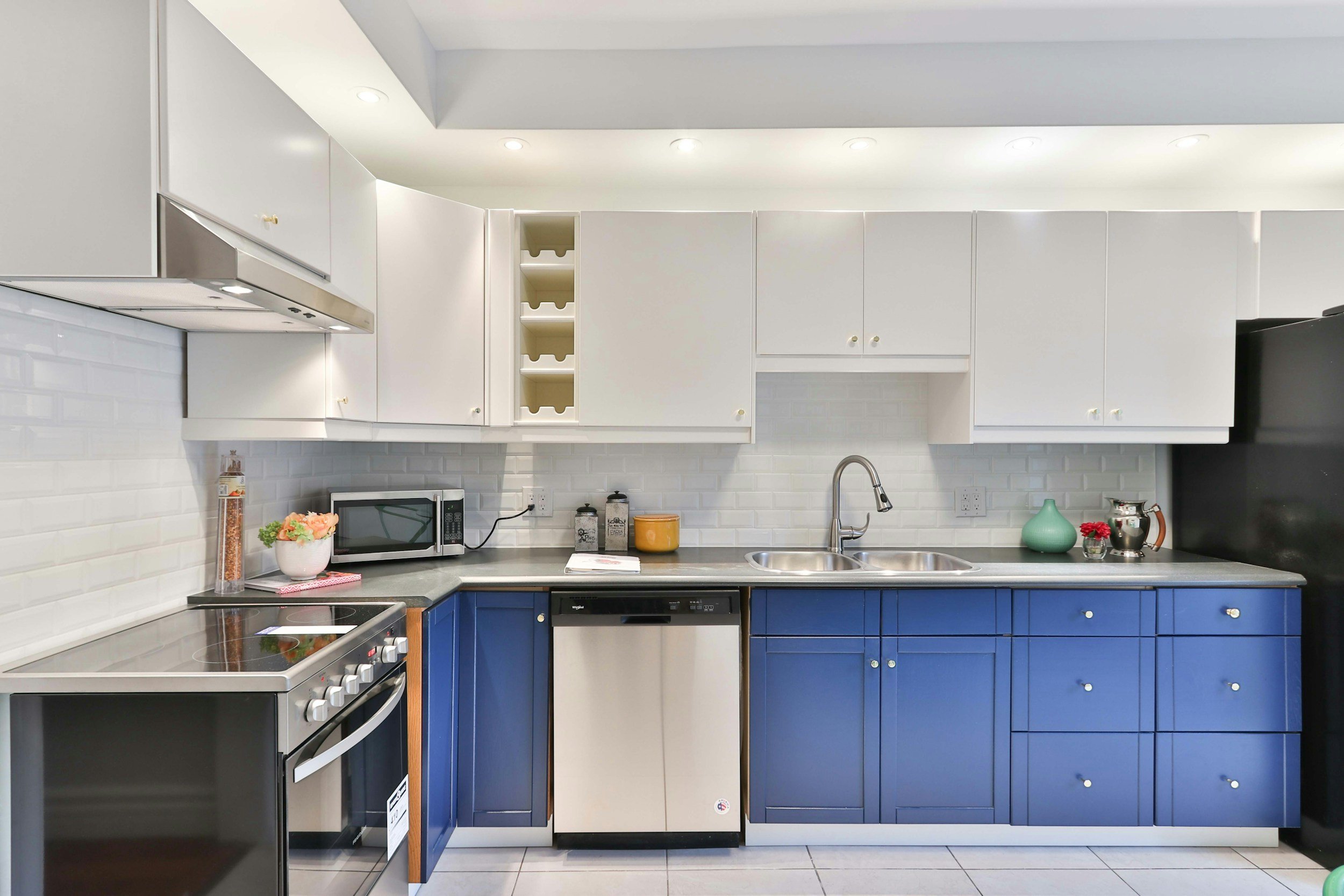 Modern kitchen with white upper cabinets, blue lower cabinets, stainless steel appliances including a stove, dishwasher, and black refrigerator, white subway tile backsplash, gray countertops, and various decorative items including a microwave, vases