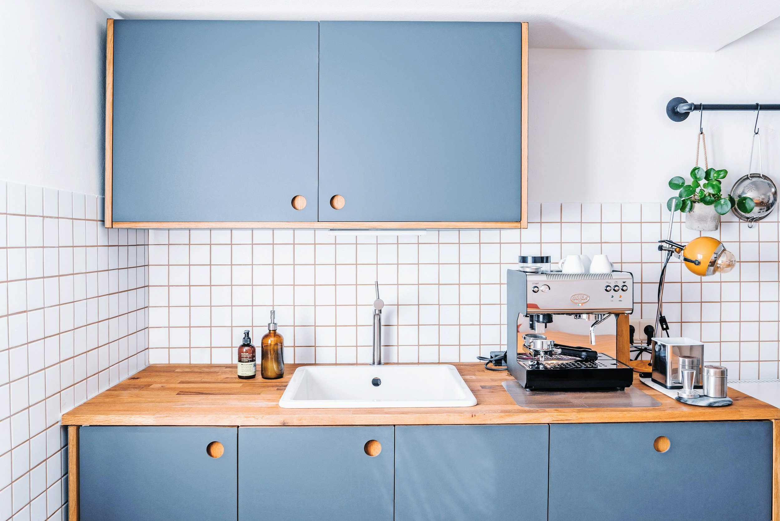 Modern kitchen with a wooden countertop, blue cabinets, white tiled wall, coffee machine on the right, a small plant, and hanging utensils. DentonPaint.Com is your go to painter in Denton County and surrounding areas for your kitchen cabinets.