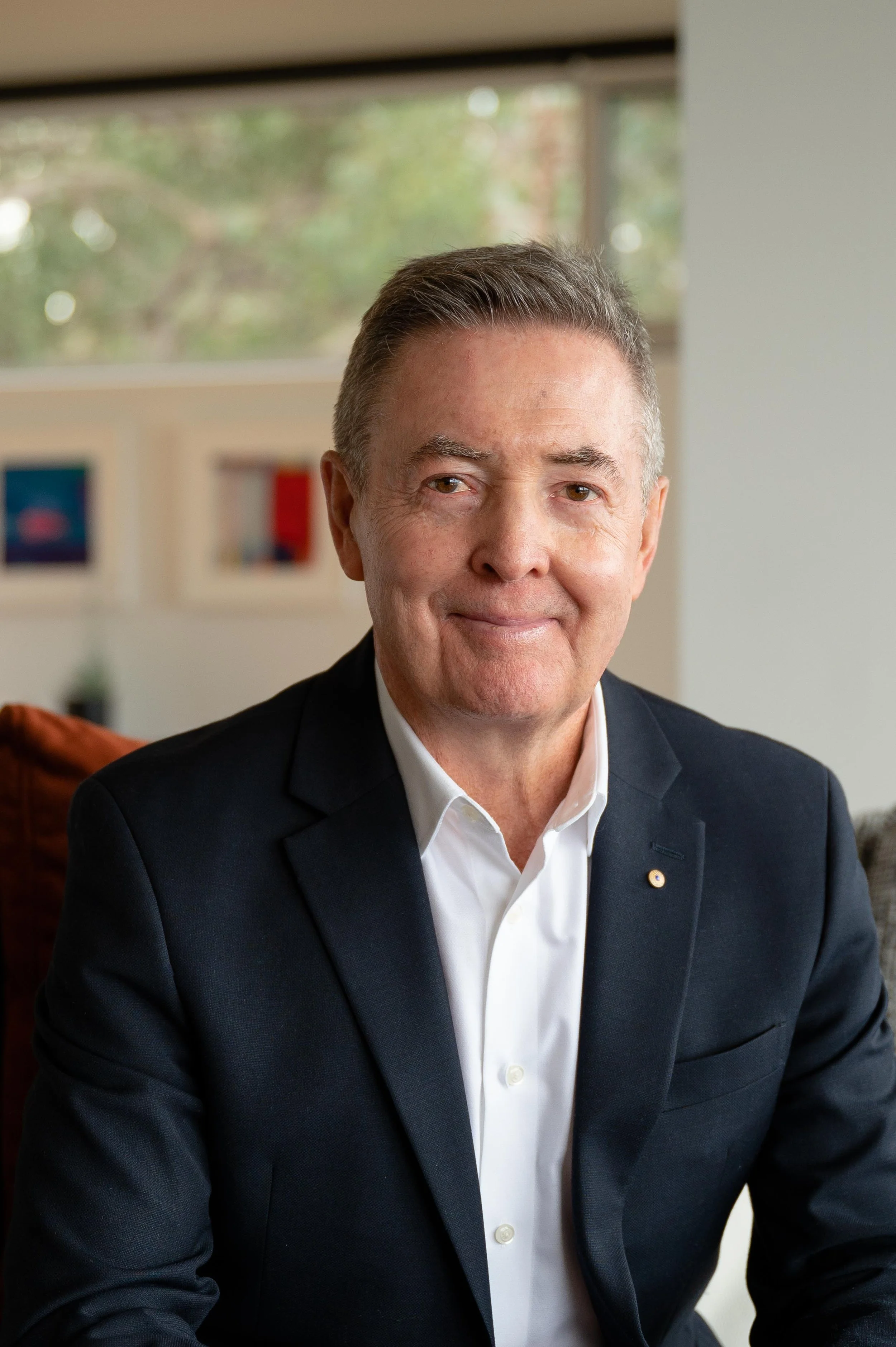 A middle-aged man in a black suit and white shirt sitting indoors with a friendly smile.