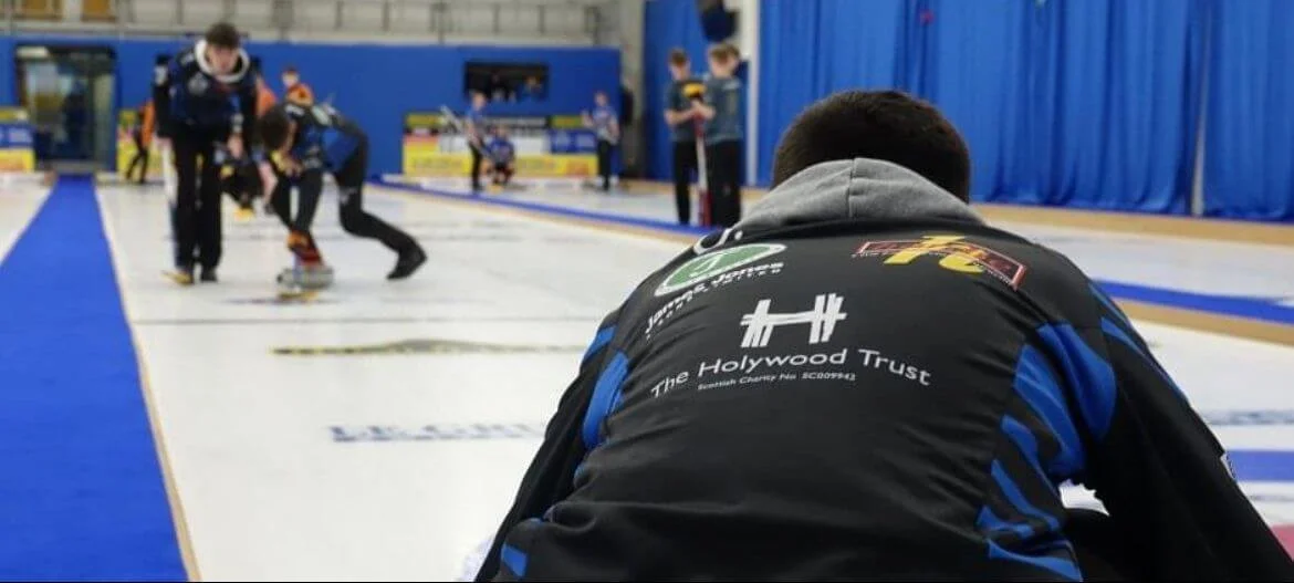 A person sitting on the ground watching young children curl at an indoor curling rink.