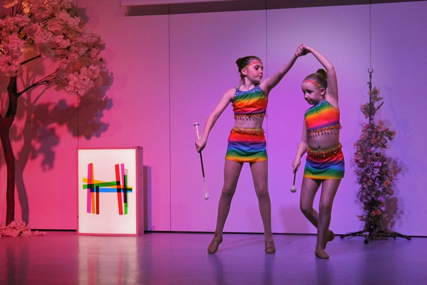 Two young girls performing a dance routine on stage, wearing rainbow-colored costumes and holding props, with pink lighting, a cherry blossom tree decoration, and Holywood Trust logo in the background.